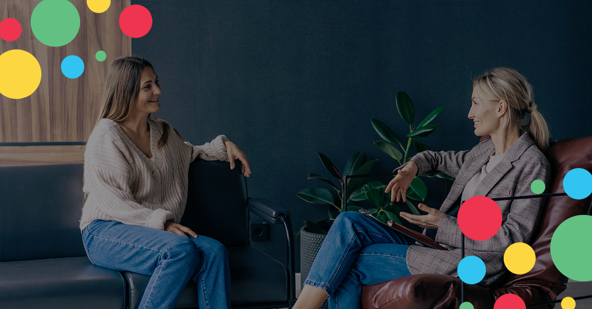 two women sitting down and talking to each other, smiling. 